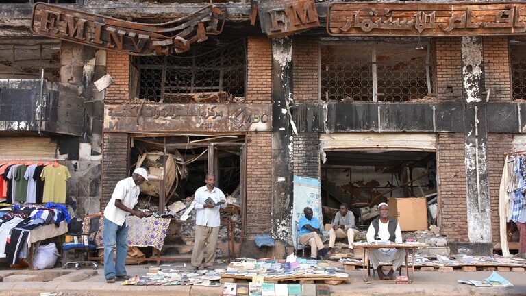 In der sudanesischen Hauptstadt Khartum herrscht auf der Straße wieder Alltag: Händler verkaufen Kleidung und Bücher, hinter ihnen steht ein Gebäude mit schwer beschädigter Fassade.