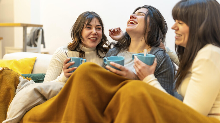 Glückliche Freundinnen, die zu Hause einen gemütlichen Moment zu dritt genießen, lachen und heiße Getränke trinken, während sie auf der Couch unter einer warmen Decke sitzen