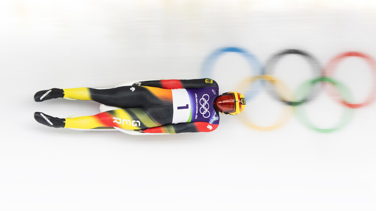 Der deutsche Athlet Max Langenhan liegt auf dem Rücken in seinem Rennrodel, während der die Rodelbahn entlang fährt. Auf der Rodelbahn sind die fünf olympischen Ringe sichtbar.
