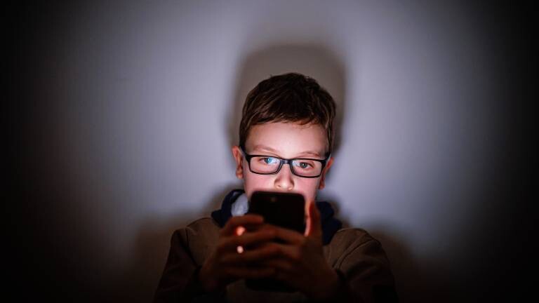 Ein Junge sitzt in einem dunklen Raum und wird von einem Smartphone beleuchtet. Symbolfoto. Gestellte Aufnahme zum Thema Kinder nutzen Social Media.