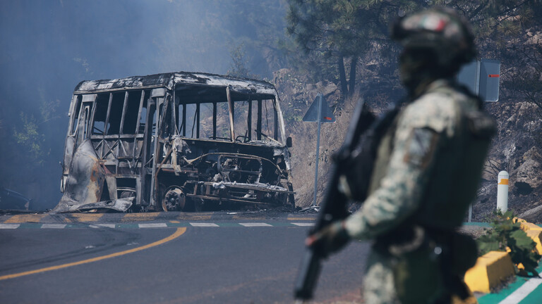 Auf einer Straße in Mexiko liegt ein ausgebrannter Bus. Davor steht ein Soldat, der eine Waffe in der hält.
