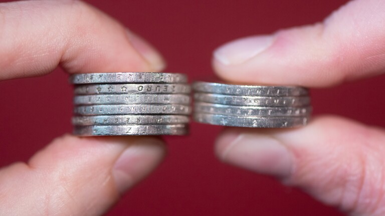 Finger halten gestapelte Zwei-Euro-Münzen nebeneinander, links sind es mehr Münzen