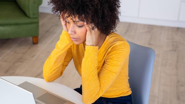 Eine Frau trägt im Homeoffice gelben Pulli und schwarze Hose. Sie sitzt am Tisch, vor ihr steht der Laptop. Sie schließt die Augen und massiert sich mit den Händen die Schläfen.