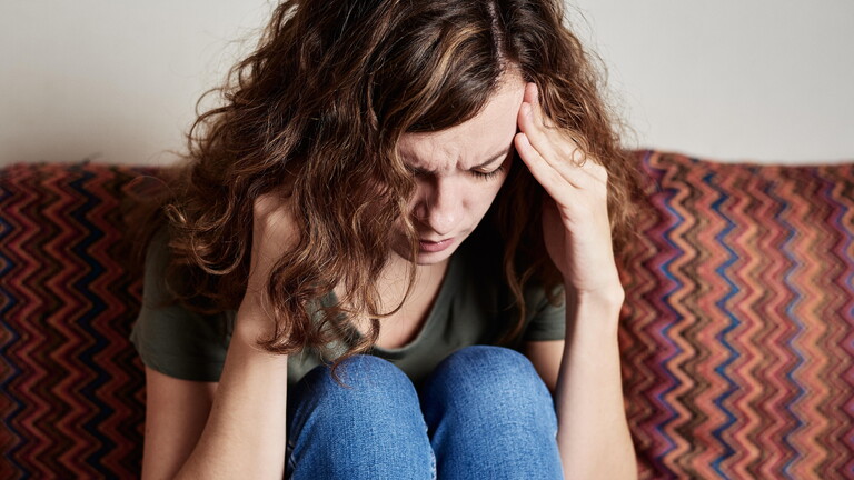 Eine junge Frau sitzt mit angezogenen Beinen auf der Couch, fasst sich an die Schläfen und kräuselt angestrengt die Stirn.