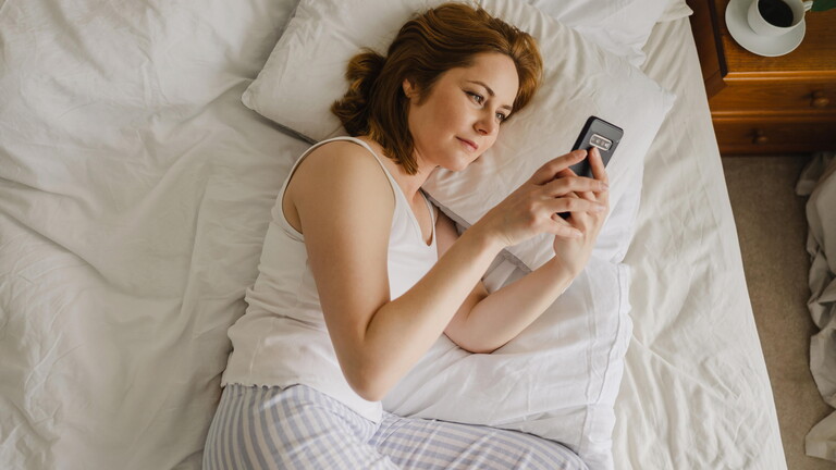 Eine Frau liegt im Bett und schaut auf ihr Handy.