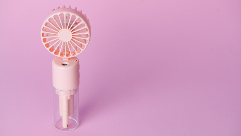 Ein kleines Ventilatorgerät mit einem Nebelsprüher steht auf einer pinken Oberfläche.