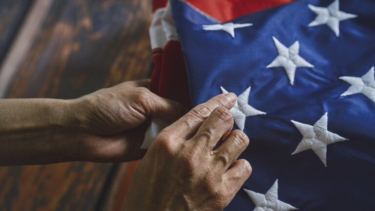 Nahaufnahme von Händen, die ehrfürchtig eine amerikanische Flagge falten und dabei komplizierte Nähte und lebendige Farben betonen. Das Bild vermittelt Respekt, Patriotismus und Liebe zum Detail