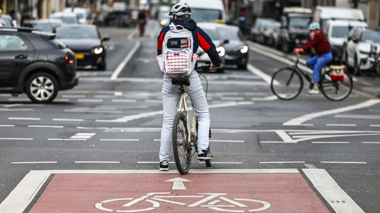 Ein Radfahrer steht auf einer Radspur an einer viel befahrenen Kreuzung. Vor ihm kreuzen Autos die Straße.