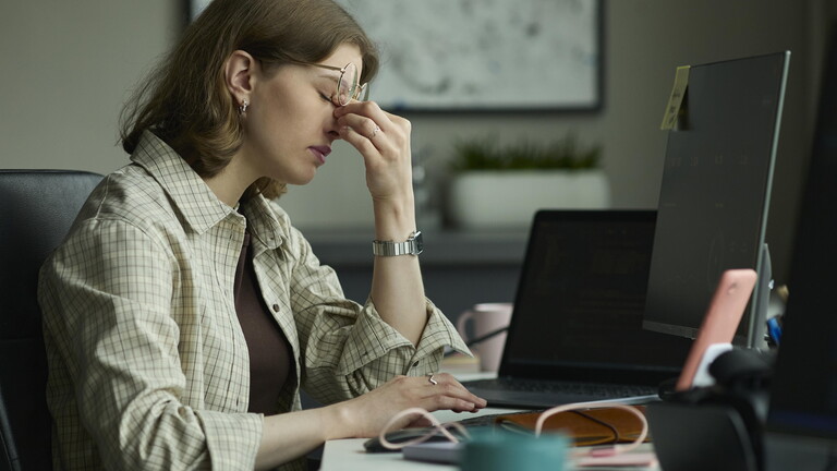 Eine junge Frau sitzt an ihrem Arbeitsplatz vor einem Computer. Sie wirkt müde und drückt mit zwei Fingern auf ihre Nasenwurzel. Dabei rutscht ihre Brille hoch.