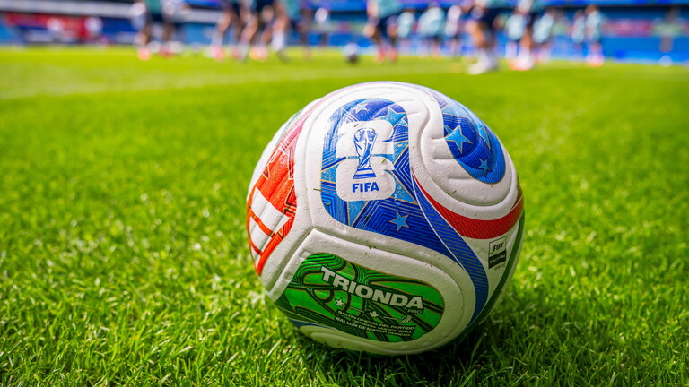 Der offizielle FIFA-Weltmeisterschaftsfußball 2026 liegt auf einem Rasen. Im Hintergrund sind Fußballspieler auf dem Fußballfeld und kicken einen Ball.
