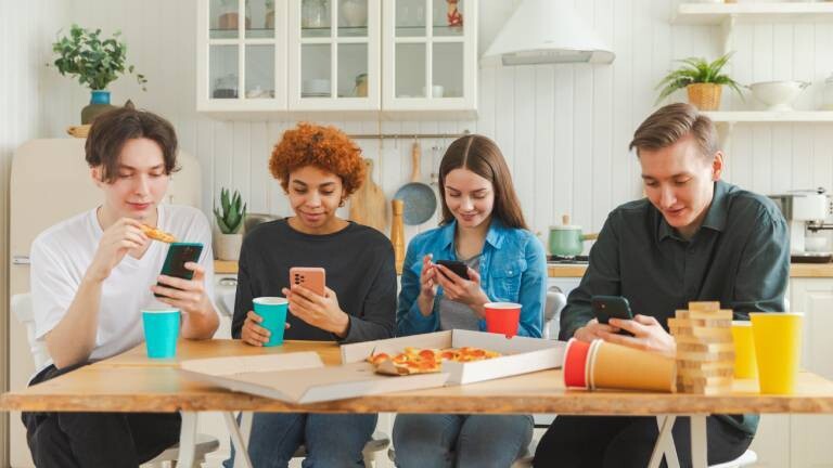 Eine Gruppe junger Leute sitzt in der Küche am Tisch. Alle gucken auf ihr Smartphone und essen bzw. trinken parallel.