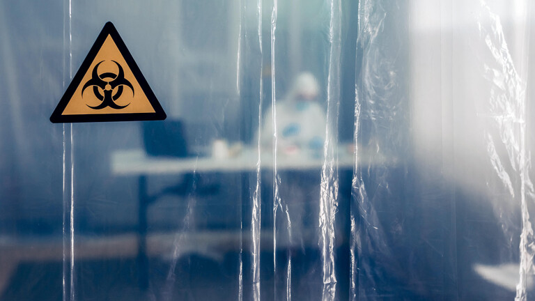 Eine Person sitzt in Schutzkleidung an einem Schreibtisch. Davor ist eine Plastikfolie angebracht mit dem weltweiten Warnsymbol für Biogefährdung.