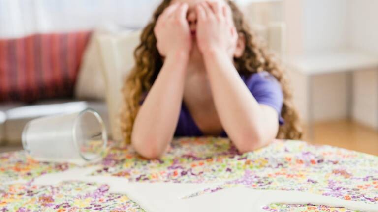 Mädchen hält sich die Augen zu. Sie sitzt an einem Tisch, auf dem ein Glas Milch verschüttet ist