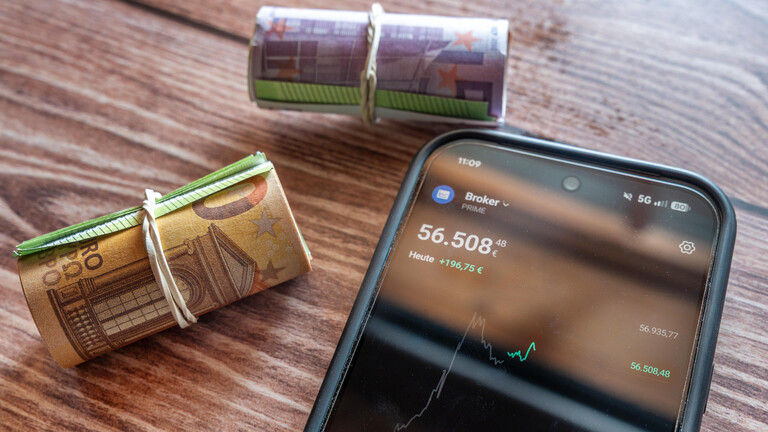 Auf einem Holztisch liegen zwei gerollte Bündel Geldscheine, die mit einem Gummi zusammengehalten werden. Daneben liegt ein Smartphone, auf dem Daten zu einem Fondguthaben angezeigt werden.