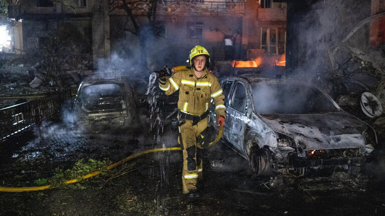 Nach einem russischen Raketenangriff in Kiew versucht eine Feuerwehrmann Brandherde zu löschen. Neben ihm stehen ausgebrannte Autos. Im Hintergrund lodern Feuer in einem Wohnhaus.