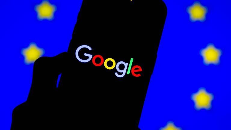 Nahaufnahme eines Smartphones, das das Google-Logo zeigt, positioniert vor einer unscharfen Flagge der Europäischen Union (EU) im Hintergrund.