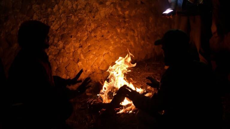 Menschen sitzen um ein Feuer verteilt und wärmen sich.
