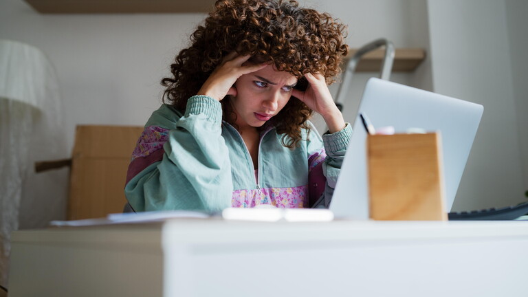 Eine Frau sitzt inmitten von Umzugskartons an einem Schreibtisch. Sie schaut auf ihren Laptop, hat ihren Kopf auf ihre Hände gestützt und wirkt gestresst.