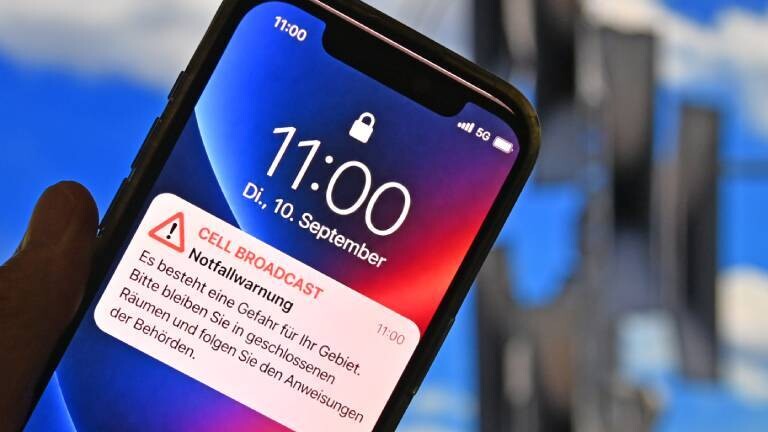 Ein Smartphone zeigt am Dienstag, 10. September um 11:00 Uhr eine Cell-Broadcast-Notfallwarnung auf dem Sperrbildschirm. Der Text fordert dazu auf, wegen einer Gefahr im Gebiet in geschlossenen Räumen zu bleiben. Im unscharfen Hintergrund ist ein Mobilfunkmast vor blauem Himmel zu sehen.