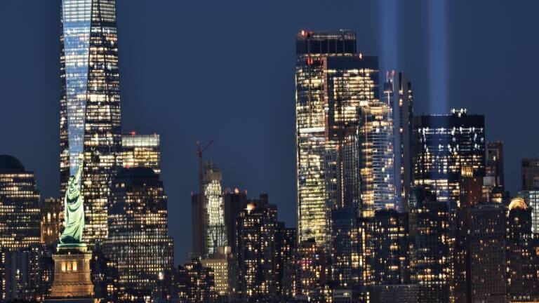 Nächtliche Skyline von New York City mit der beleuchteten Freiheitsstatue links und den zwei hellblauen Lichtsäulen des "Tribute in Light" im Hintergrund.