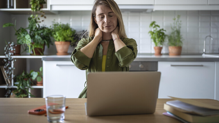 Eine Frau sitzt an einem Küchentisch am Laptop. Sie hält ihre Hände an ihrem Nacken und hat dabei die Augen geschlossen.