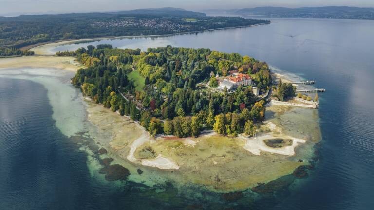 Eine Luftaufnahme zeigt die Insel Mainau im Bodensee bei extremem Niedrigwasser aufgrund des Klimawandels. Die sonst tiefblaue Uferzone liegt weitflächig trocken und gibt helle Sand- und Kiesbänke sowie grünliche Algen- und Sedimentstrukturen unter der flachen Wasseroberfläche frei.