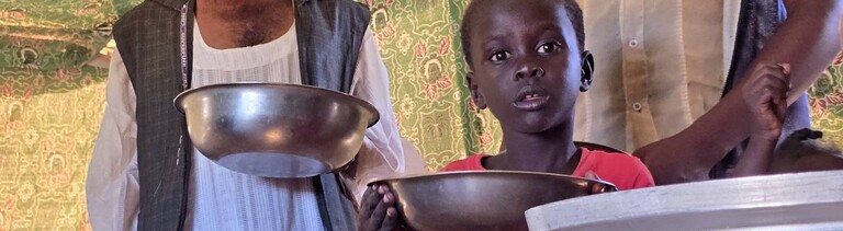 Ein Mann und ein Kind stehen mit Schüsseln in der Hand an einer Essensausgabe (Meroë/Sudan, 09.01.2025)