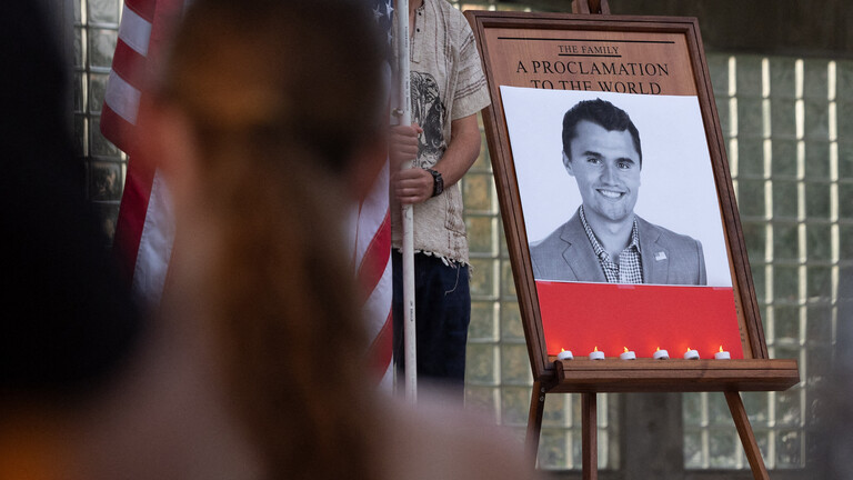 Ein Schwarz-Weiß-Foto on Charlie Kirk bei einer Trauerfeier, daneben die US-Flagge, im Vordergrund Menschen
