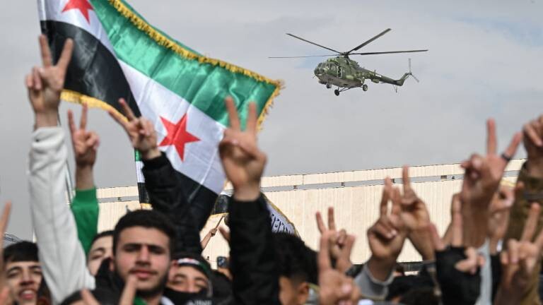 Ein Hubschrauber fliegt über eine Menschenmenge. Das Siegeszeichen und Flaggen werden gezeigt.  Es handelt sich um Feierlichkeiten zum Jahrestag des Sturzes des Machthabers Baschar al-Assad in der syrischen Hauptstadt Damaskus (8.12.2025, Syrien)