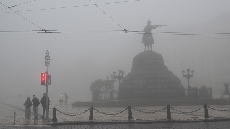 Menschen stehen in dichtem Nebel an einer AMpel in Kiew, im Hintergrund ein Militär-Denkmal.