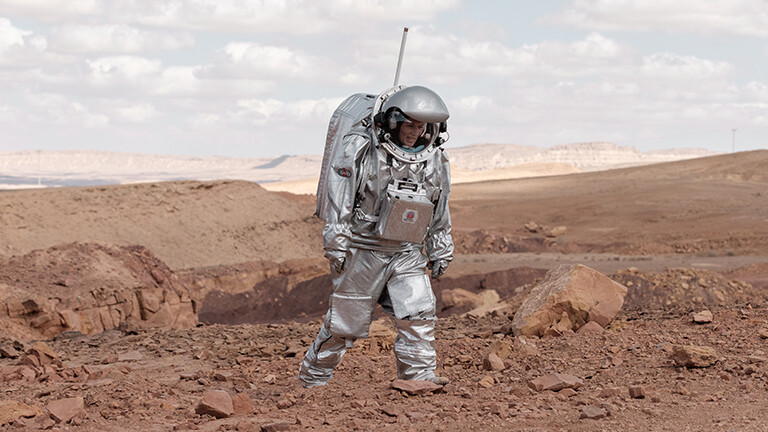 Foto der Analog-Astronautin Anika Mehlis, die in einem schweren, silbernen Raumanzug-Simulator im Rahmen einer Marsmission-Simulation durch die Wüste stapft