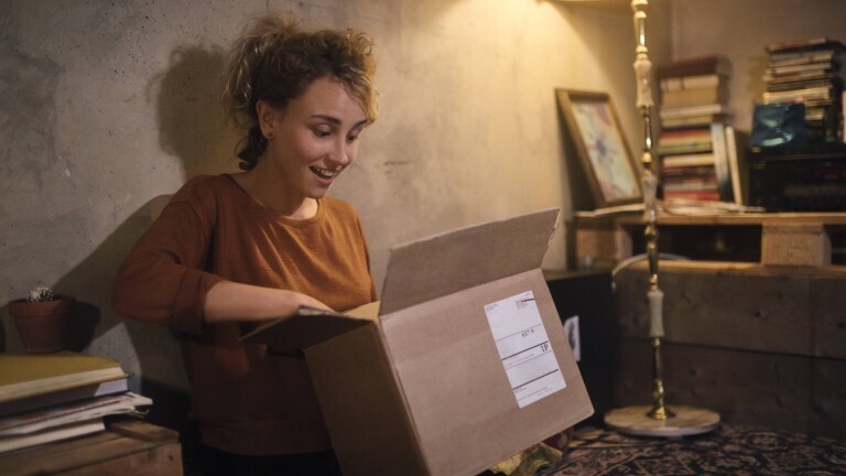 Eine Frau packt freudig etwas aus einem Karton aus. Sie sitzt am Boden. Im Hintergrund leuchtet eine Steglampe und es ist ein Bücherstapel zu sehen.