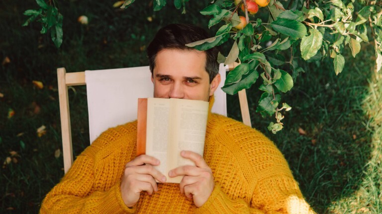 Ein Mann sitzt draußen in einem Liegestuhl unter einem Apfelbaum. Er hält sich ein Buch vor den Mund. Die Hälfte seines Gesichts ist dadruch verdeckt.