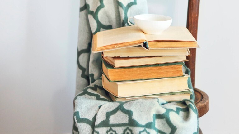Auf einem Holzstuhl liegt eine Decke. Darauf stapeln sich Bücher. Oben auf den Büchern ist eine Teetasse.