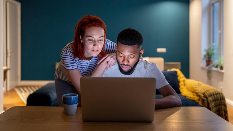 Ein Mann sitzt vor einem Laptop. Eine Frau lehnt sich an seine Schulter und schaut gemeinsam mit ihm auf den Computer.