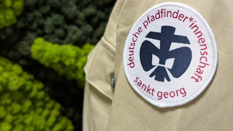 Das Logo der deutschen Pfadfinder*innenschaft Sankt Georg auf einem beigefarbenen Pfadfinderhemd