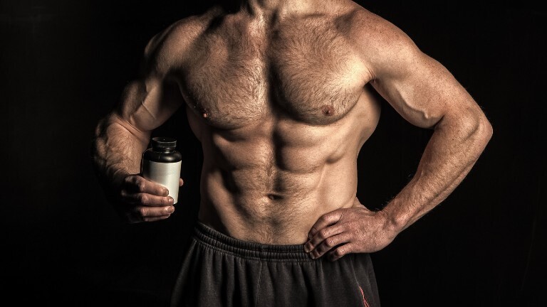 Im Bild ist ein muskulöser Männeroberkörper zu sehen. Der Mann hält in einer Hand ein Glas, das an ein Medikamentenbehälter erinnert.