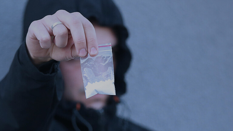 Gestelltes Foto eines Drogendealers der ein Tütchen mit einem weißen Pulver in die Kamera hält.