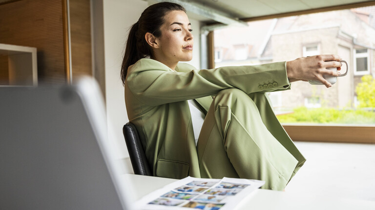 Eine Frau sitzt in einem grünen Anzug in einem Büro auf einem Stuhl, hält dabei eine Tasse in der Hand und wirkt nachdenklich.