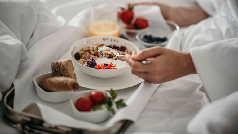 Eine Person frühstückt im Bett. Es ist ein Tablett mit Brötchen, Erdbeeren, Jogurt und Orangensaft zu sehen.