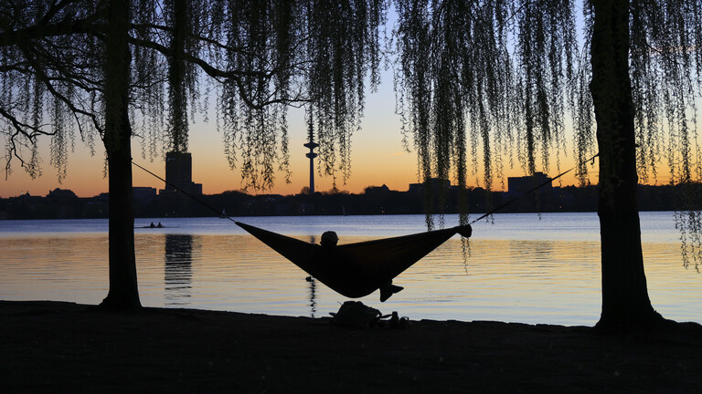 Ein Mensch liegt an einem See in einer Hängematte. Im Hintergrund sind Gebäude erkennbar. Die Sonne geht gerade unter.