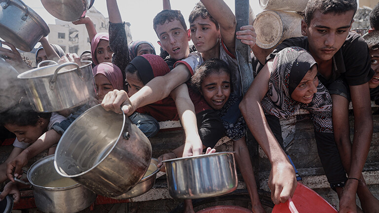 Foto einer dichtgedrängten Menschenmenge im Gazastreifen, darunter Kinder, die teils verzweifelt  aussehend leere Töpfe und Gefäße in Richtung der Kamera strecken um Lebensmittel entgegenzunehmen, die von einer Wohltätigkeitsorganisation verteilt werden (4. August 2025)