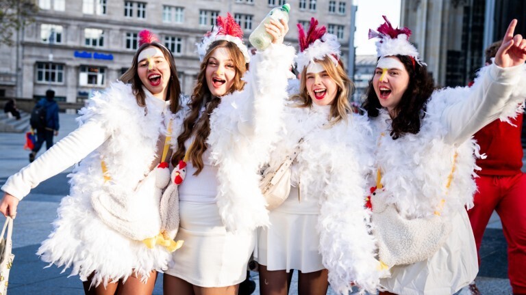 Vier junge Frauen haben sich an Karneval als Hühner verkleidet. Sie stehen nebeneinander und schauen jubelnd in die Kamera.