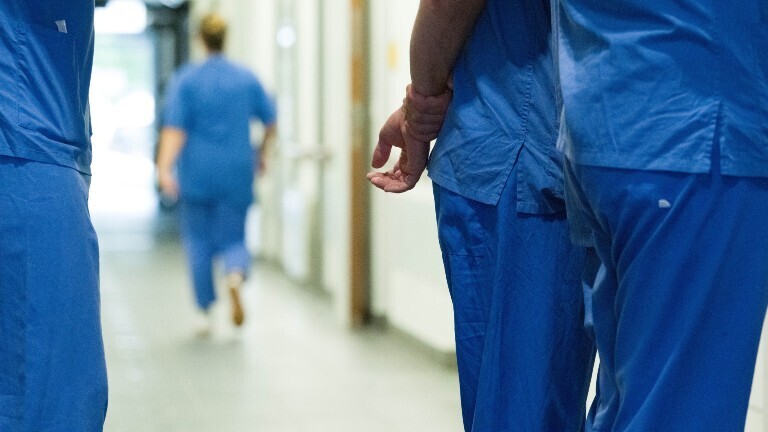 Im Gang eines Krankenhauses stehen mehrere medizinische Fachkräfte zusammen. Im Bild sieht man nur ihre Körper, aber nicht ihre Gesichter. Sie tragen blaue Arbeitskleidung.