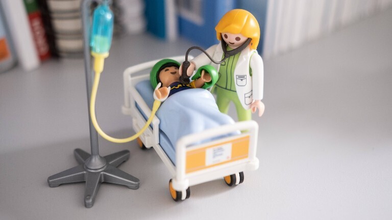 Zwei Playmobil-Figuren stehen auf einem Tisch. Sie sollen eine Ärztin darstellen, die einen Patienten in einem Krankenhausbett untersucht.