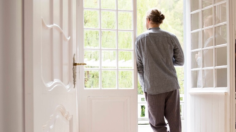 Ein Mann lehnt am Rahmen einer Balkontür. Er schaut nach draußen. Wir sehen ihn von hinten.
