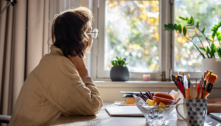Foto einer jungen Frau, die nachdenklich am Schreibtisch sitzt und aus dem Fenster guckt