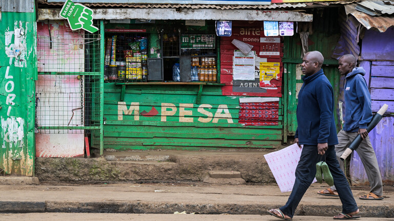 Zwei Fußgänger laufen in Kenia an einem Kiosk vorbei, auf dem für M-Pesa geworben wird.