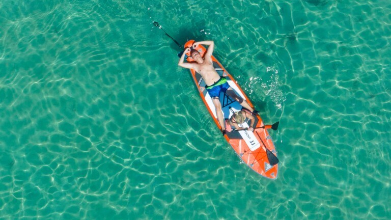 Ein Mann ruht sich auf dem Rücken liegend auf einem Stand Up Paddle aus, eine Frau sitzt auf dem Boot. Das Wasser im Hintergrund ist türkis-grün.