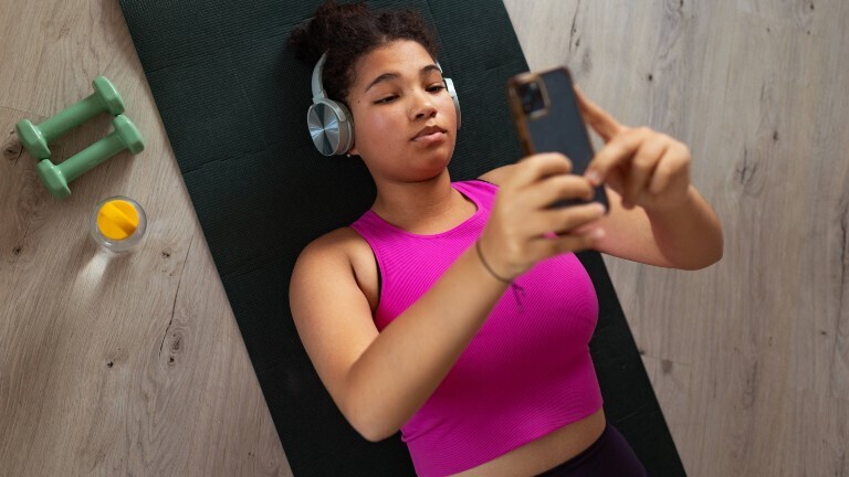 Eine junge Frau liegt auf dem Rücken auf einer Trainingsmatte und schaut dabei in ein Handy. Sie trägt Kopfhörer. Es liegen Kurzhanteln zum Muskeltraining neben ihr.